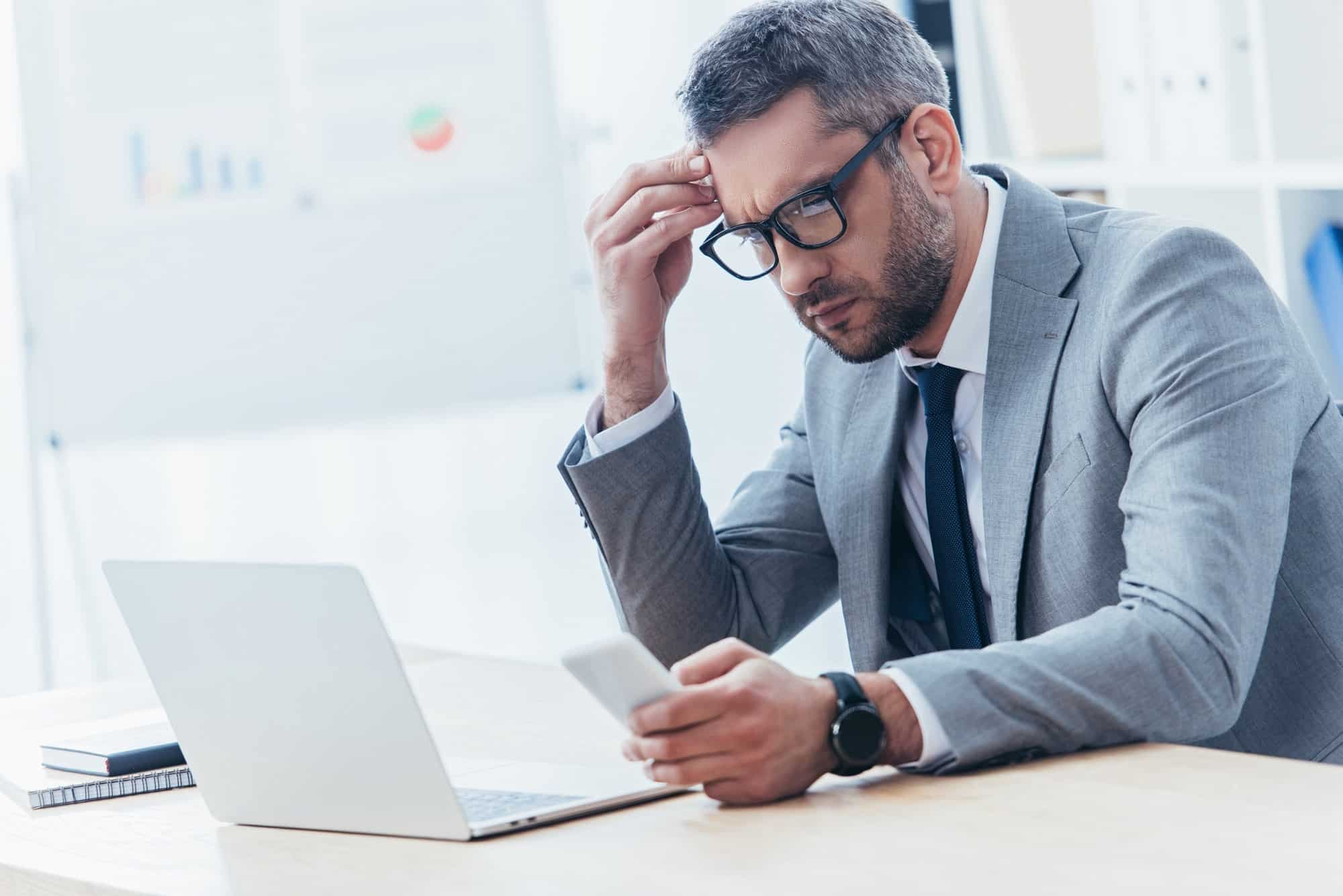 concentrated businessman in eyeglasses using smartphone and working with laptop in office