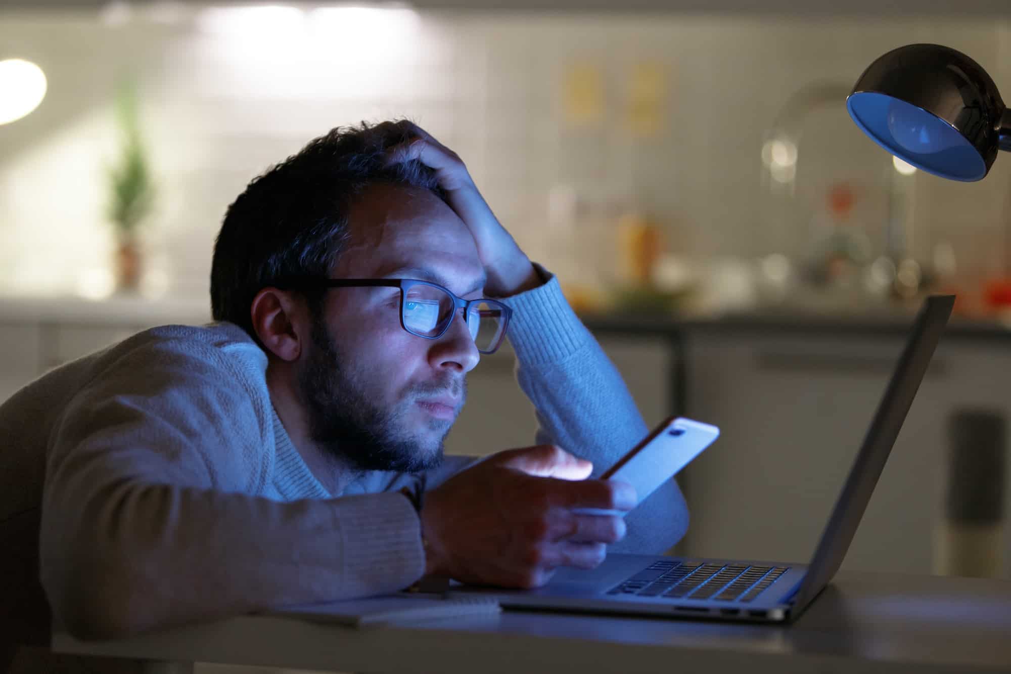 Sleepy bored man designer in eyeglasses using smartphone at night home