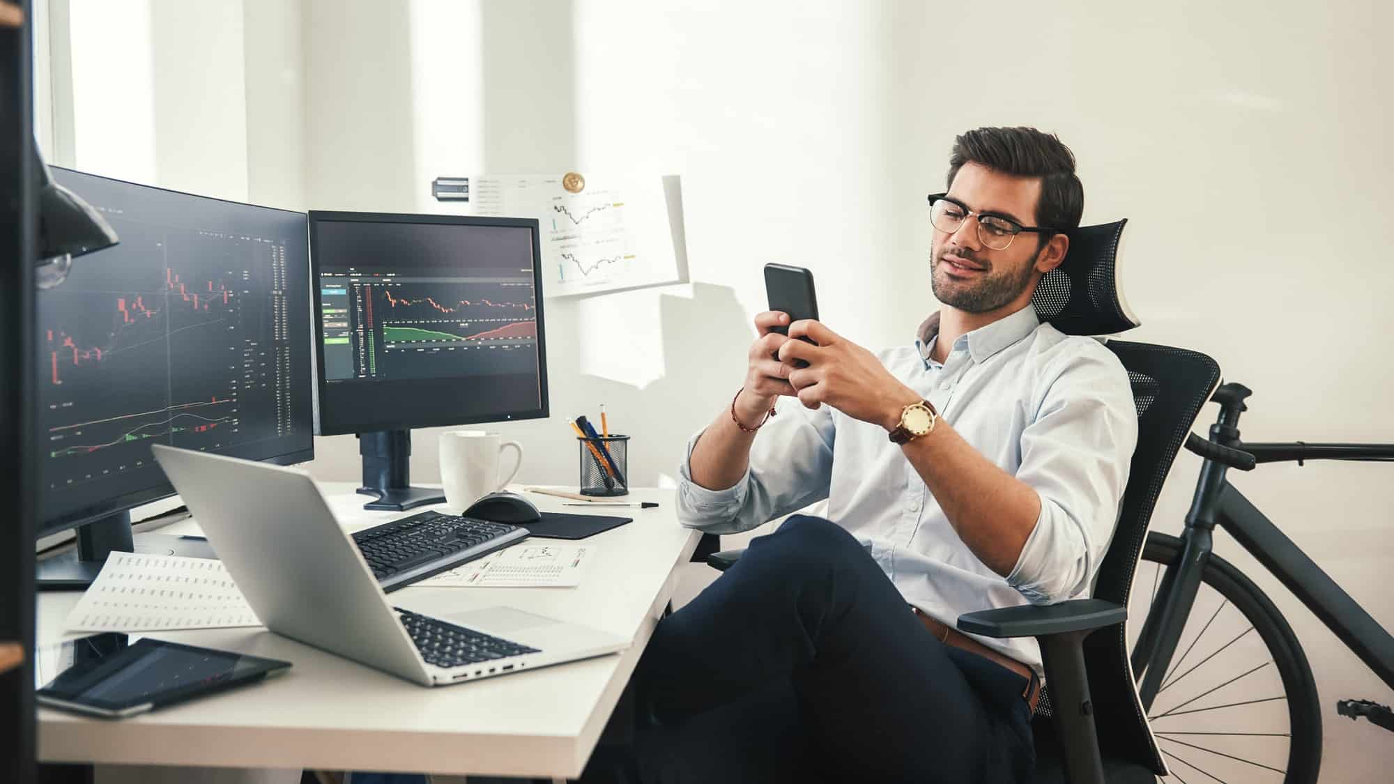 Using modern technologies. Relaxed young trader in formal wear and eyeglasses is using his